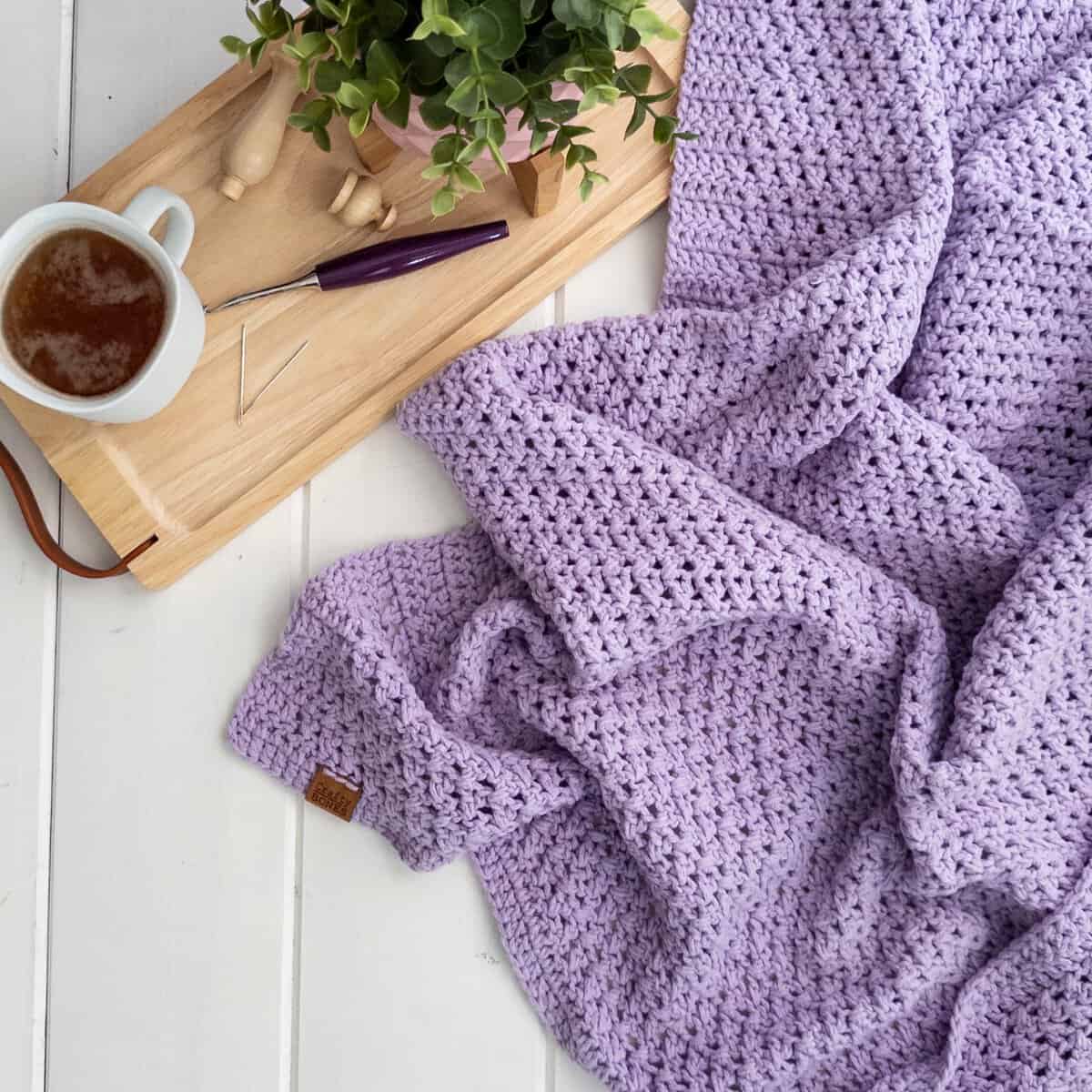 Purple Crochet blanket featuring a criss cross pattern. With a cup of tea and a furls crochet hook beside