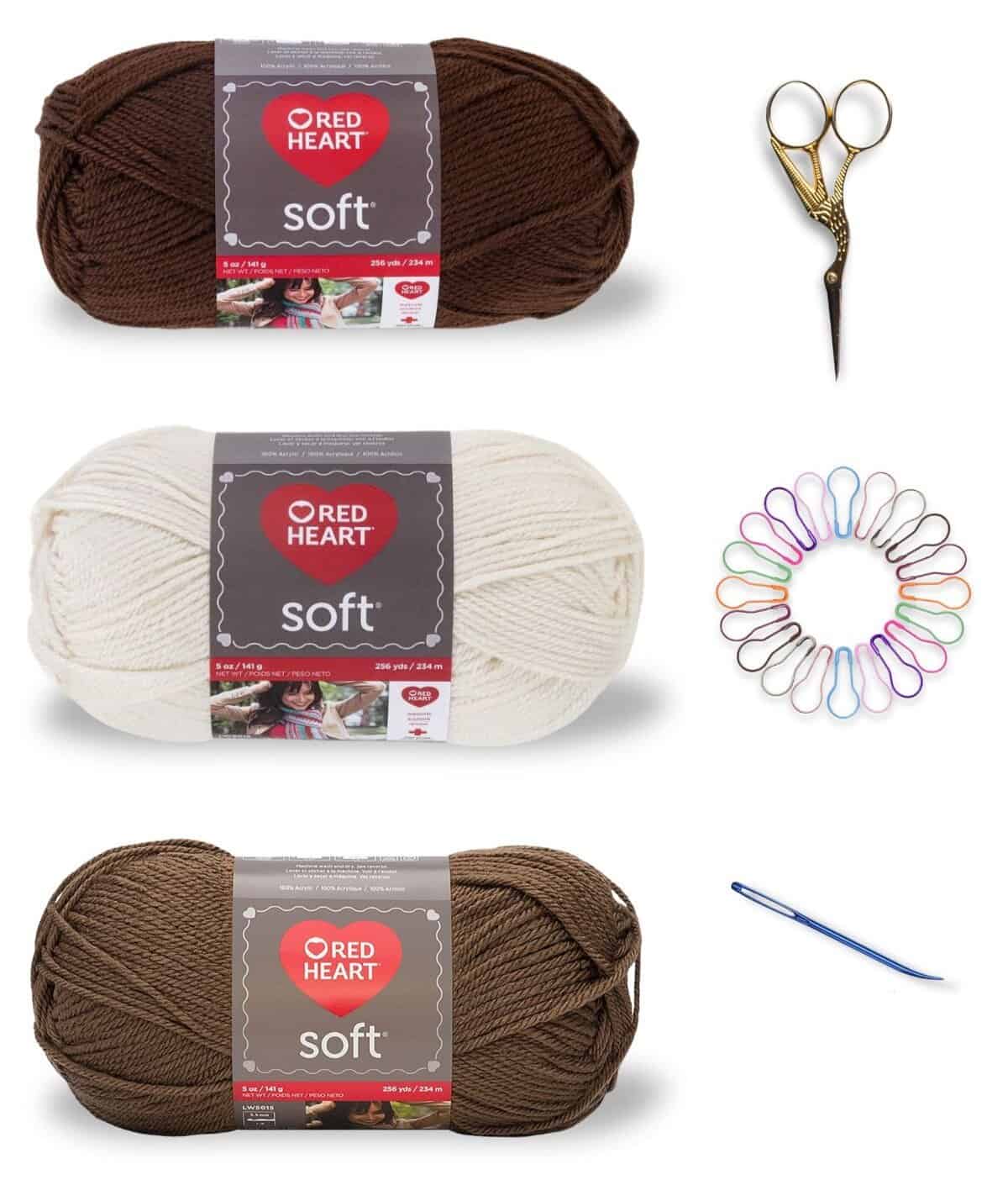 Three balls of yarn with the label red heart soft.  In a dark brown, brown, and off white colour.  A pair of gold bird shaped scissors, circle of stitch markers, and a tapestry needle.