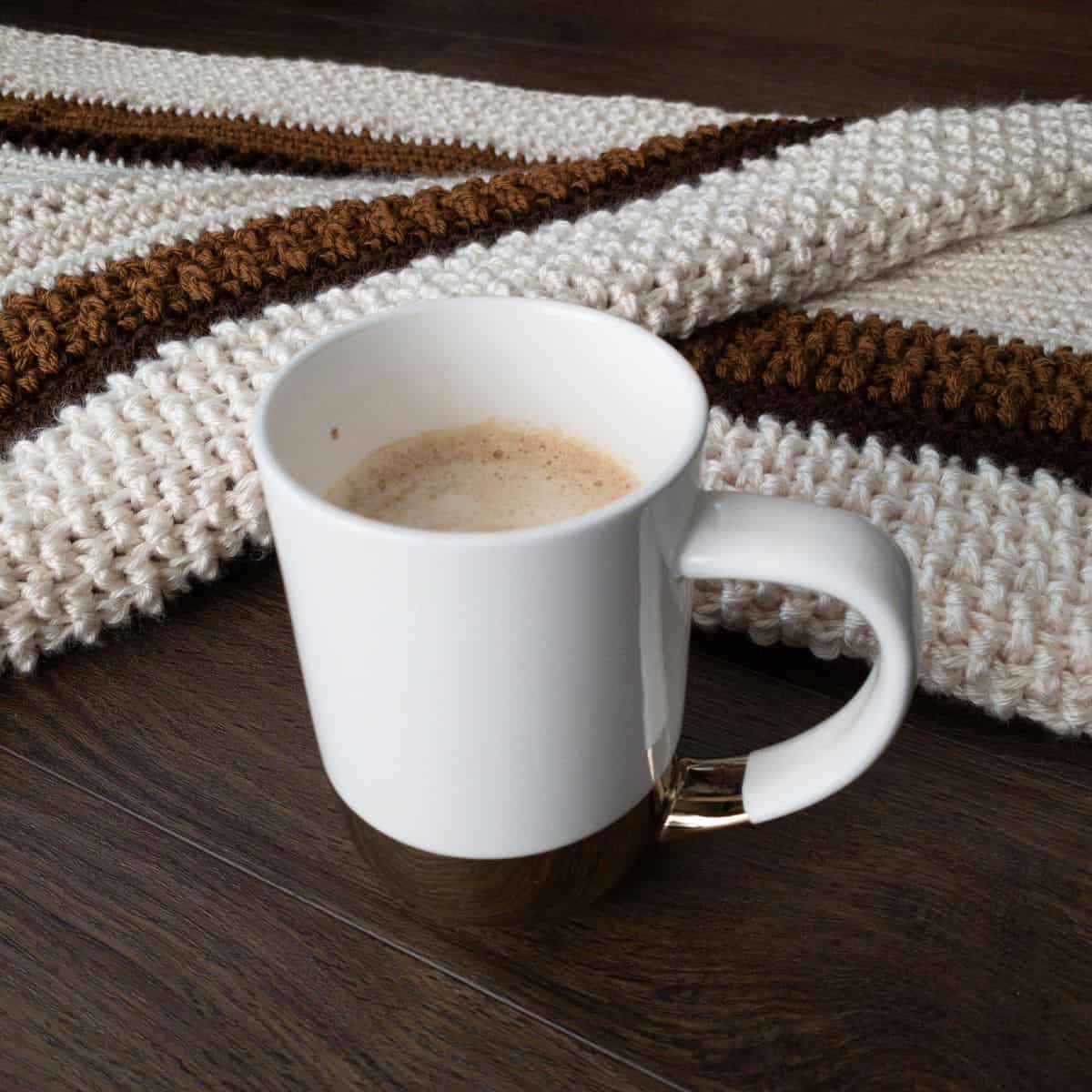White coffee mug placed in front of a cream, caramel, and chocolate brown textured crochet blanket.