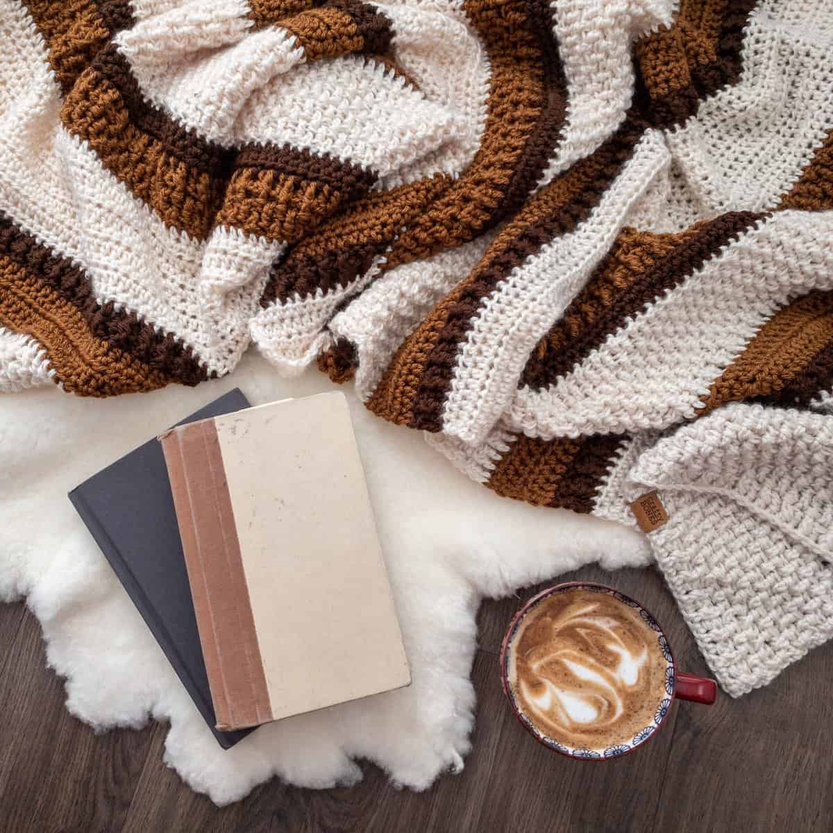 Cream and brown textured crochet blanket folded beside a cup of latte, vintage books, and a soft white rug.
