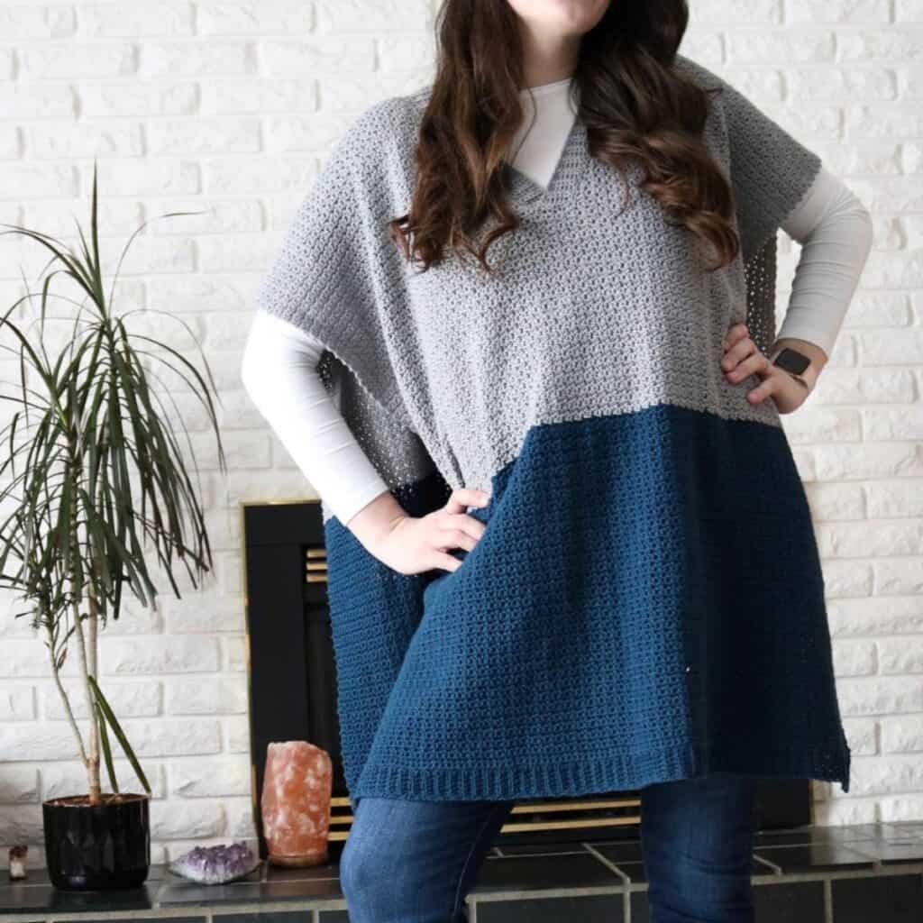 Woman modeling the Corrie Poncho, an easy beginner crochet poncho with a color-blocked design in grey and navy, standing confidently in front of a white brick fireplace.