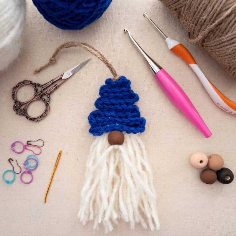 A handmade crochet gnome ornament with a blue hat, white yarn beard, and a wooden bead nose, displayed alongside crochet tools, colorful stitch markers, scissors, twine, and yarn balls. Wooden beads in different colors represent diverse skin tones, emphasizing inclusivity in the design.