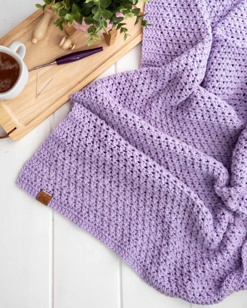 Lay flat image of the criss cross crochet blanket by crafty bones along side a tray containing a small plant, furls crochet hook, and a mug of tea