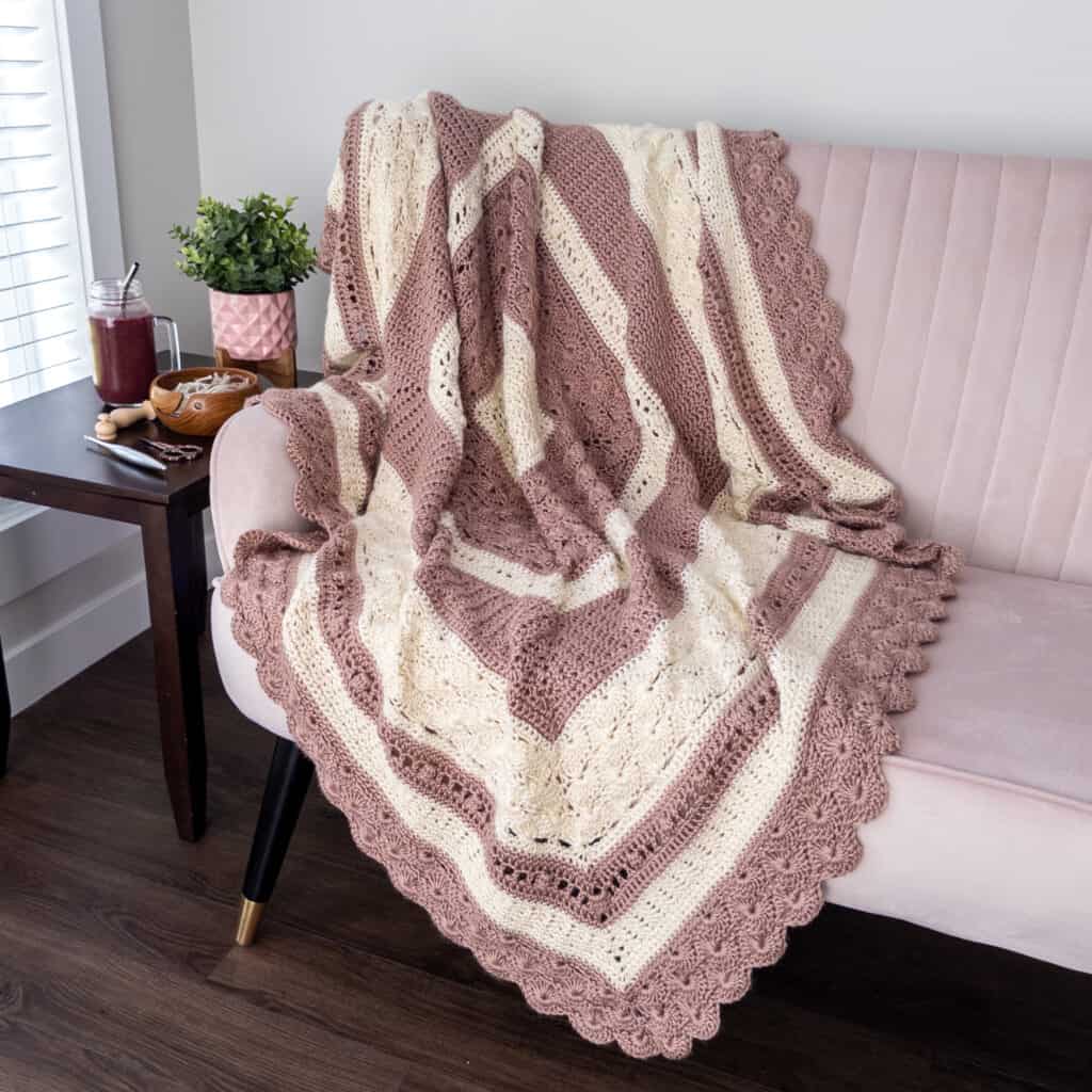 staged photo of the shell and lace heirloom crochet blanket on a light pink couch. with a smoothie, yarn bowl and crochet hook on a table beside