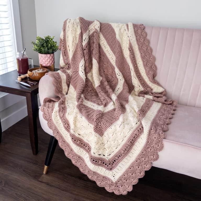 staged photo of the shell and lace crochet blanket on a light pink couch. with a smoothie, yarn bowl and crochet hook on a table beside