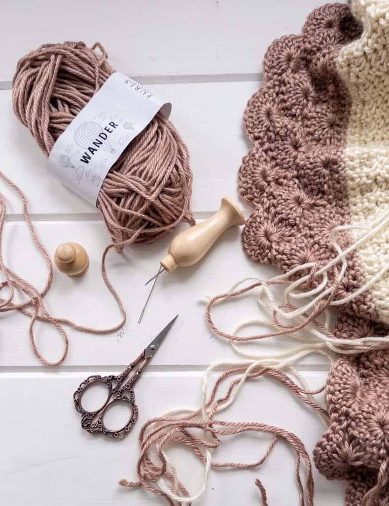 flat lay showing the shell and lace throw blanket with strings of yarn needing to be woven in, half used ball of yarn, tapestry hook and scissors