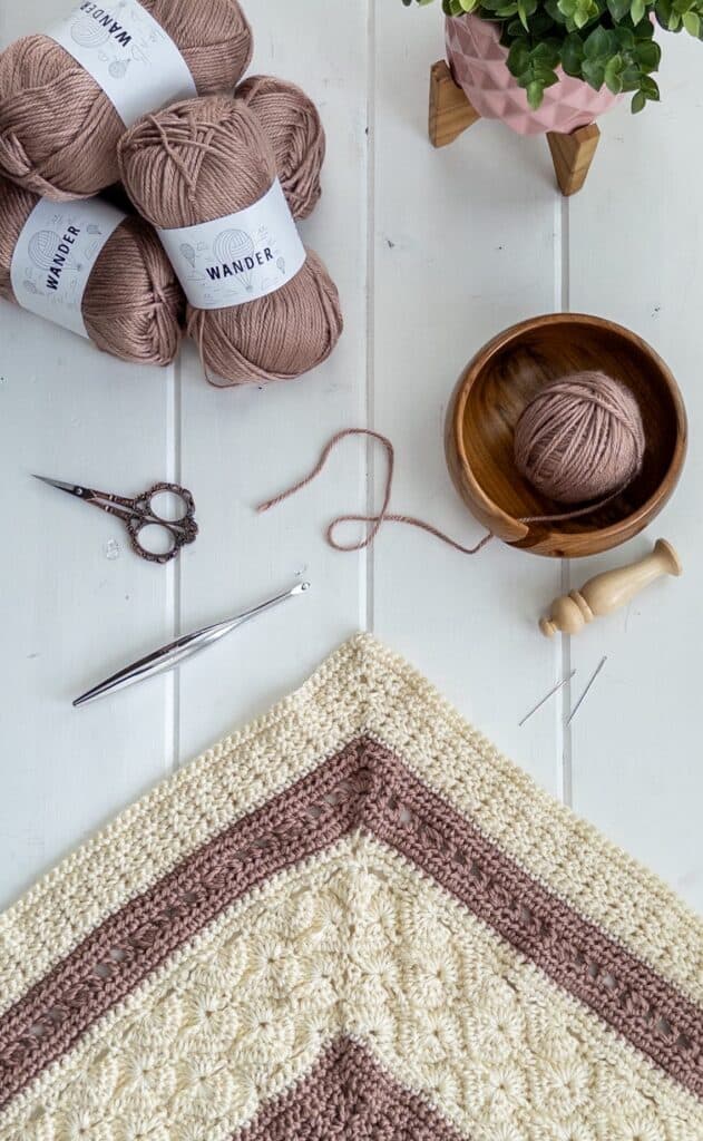 flat lay showing a stack of wander yarn, crochet hook, scissors, wood yarn bowl, and partially finished shell and lace throw