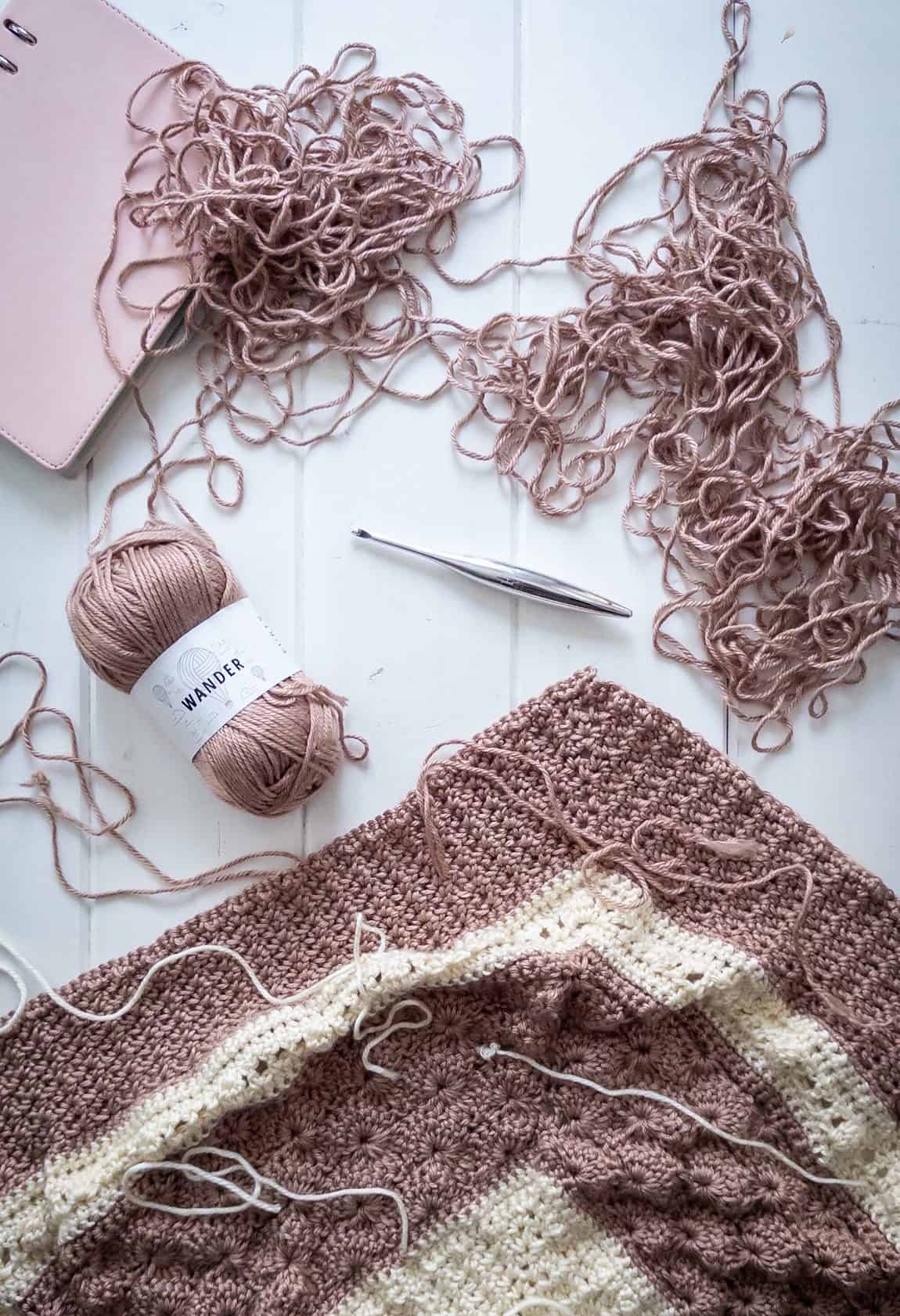Crochet blanket, yarn ball, pink binder, metal crochet hook and piles of unwound yarn