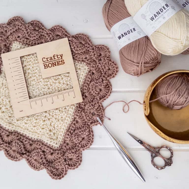 Flat lay with ball of wander yarn in two colors, a wood square measurement with the crafty bones logo, start of the shell and lace throw, crochet hook, scissors and shiny yarn bowl