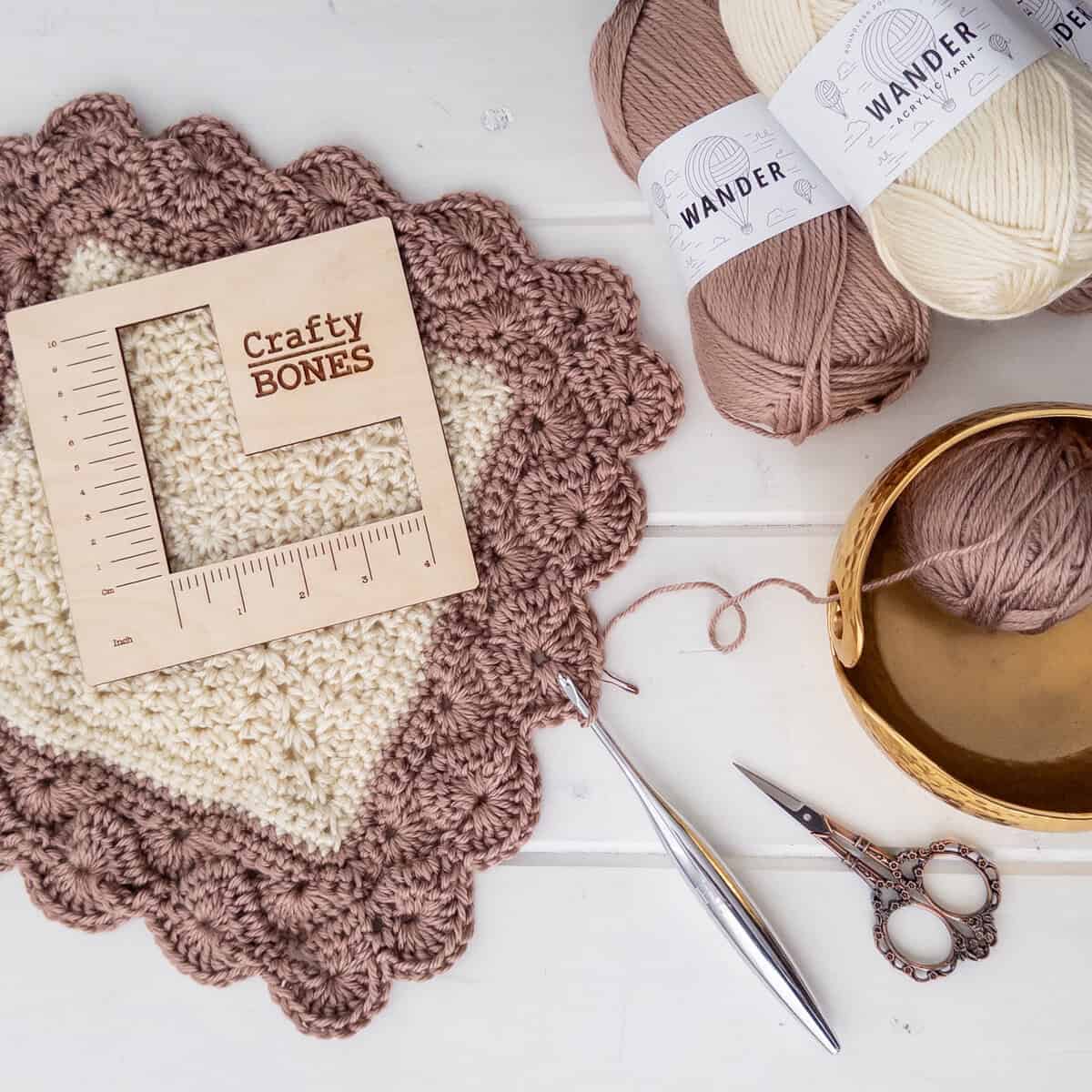 Flat lay with ball of wander yarn in two colors, a wood square measurement with the crafty bones logo, start of the shell and lace throw, crochet hook, scissors and shiny yarn bowl