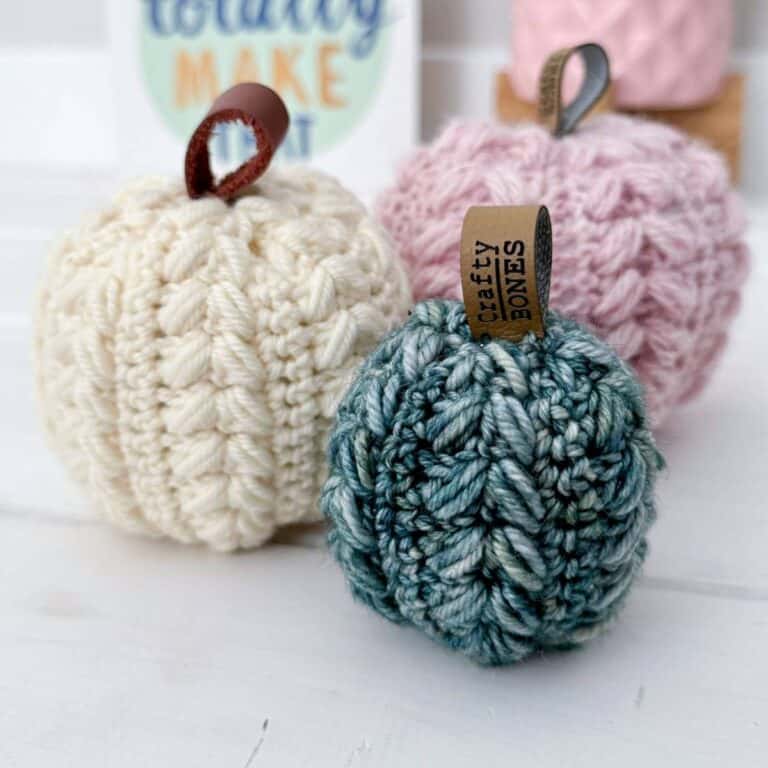 A group of three crocheted pumpkins in different colors—cream, pink, and blue-green—each with a leather or faux-leather loop stem, featuring a 'Crafty Bones' tag on the front pumpkin.