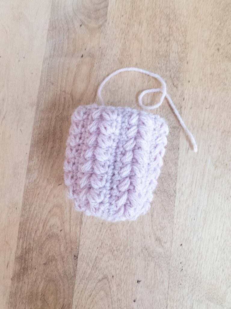 Partially finished pink crocheted pumpkin on a light wood surface, cinched on one side with the other side open for stuffing.