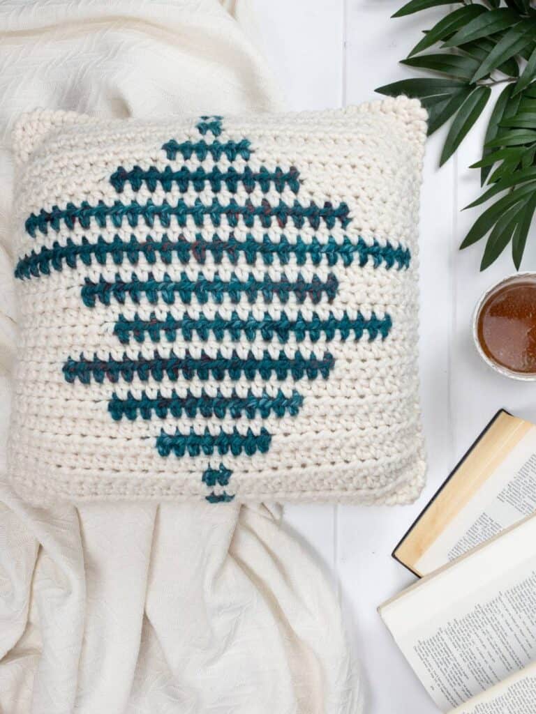 Geometric crochet pillow with teal and cream design, placed on a cozy blanket with a book and a drink.