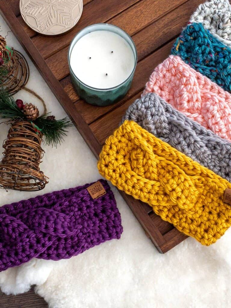 A collection of braided crochet earwarmers in vibrant colors, displayed neatly on a wooden tray alongside a candle and festive holiday decorations.