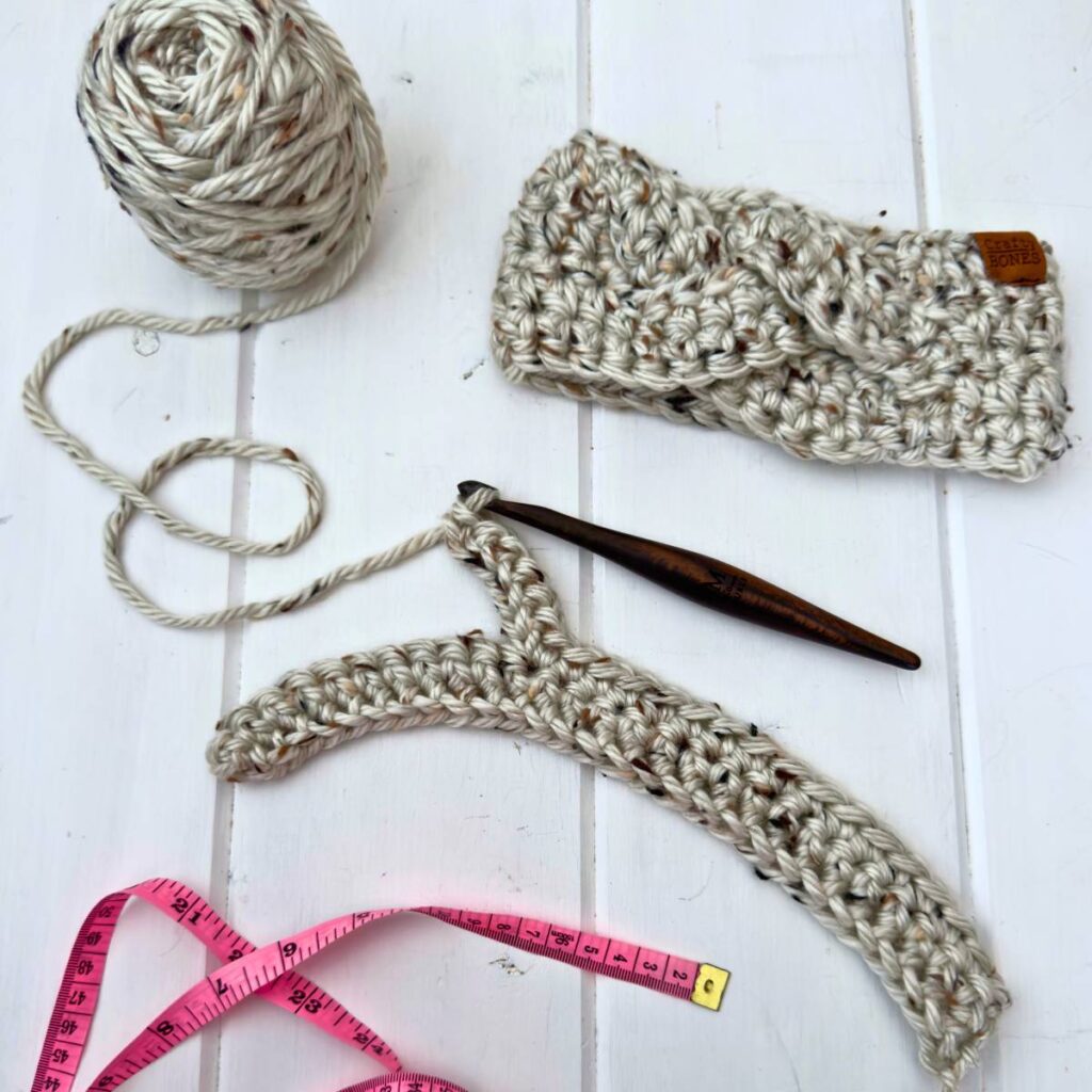 A work in progress of a crochet braided earwarmer with a skein of yarn, measuring tape, and a wooden crochet hook on a white wooden surface.