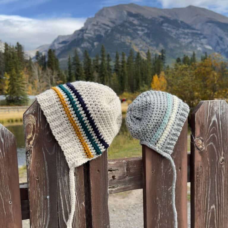 Two crochet bonnets with stripe patterns hanging on a fence with mountains in the background.