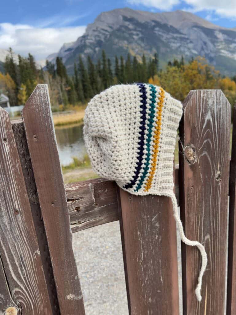 Handmade crochet bonnet with colorful stripes on a wooden fence, with mountains in the background.