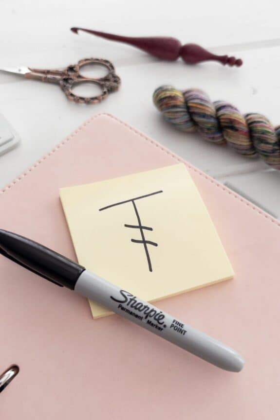 Yellow sticky note with a symbol for treble crochet on it, written in black ink on a pink and white background. Surrounded by a keyboard, crochet hook, yarn and scissors.