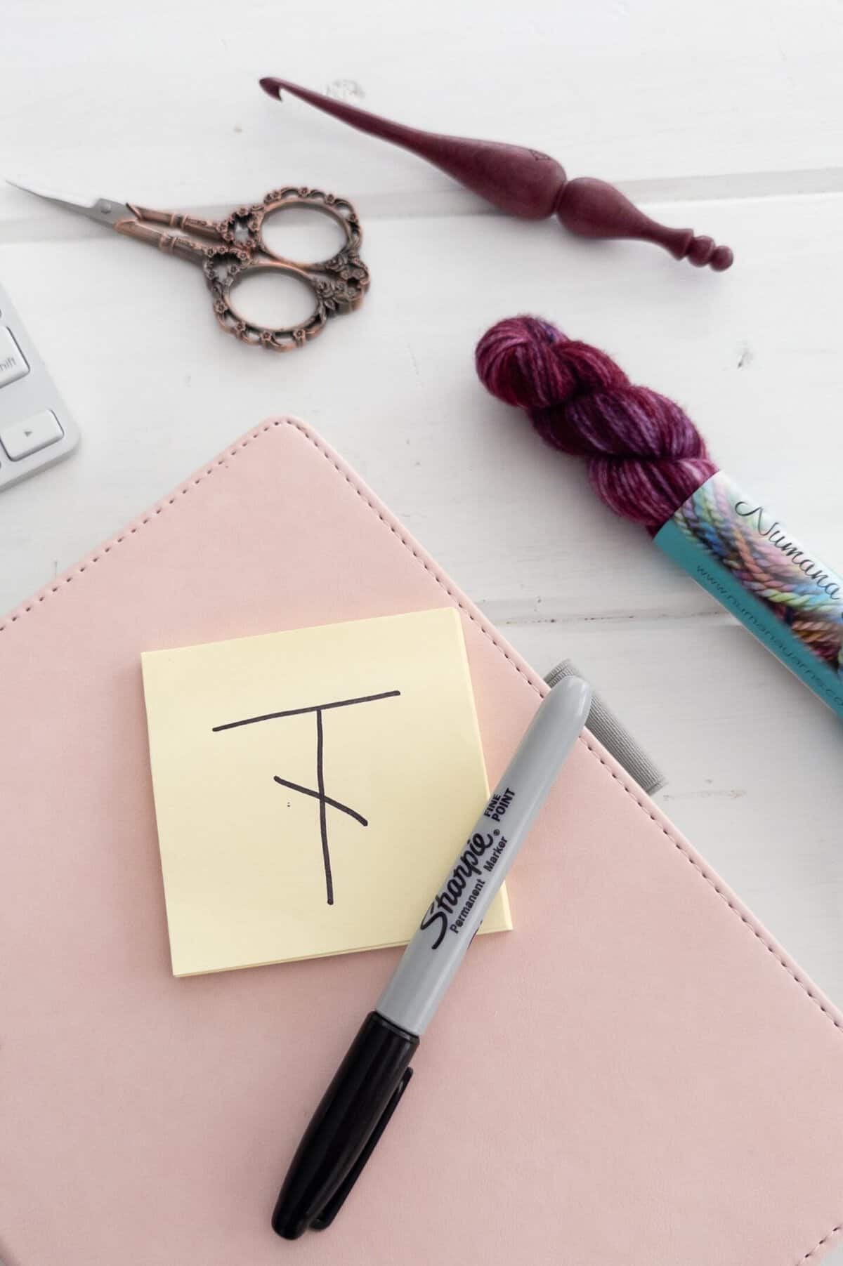 Yellow sticky note with a symbol for double crochet on it, written in black ink on a pink and white background.  Surrounded by a keyboard, crochet hook, yarn and scissors.