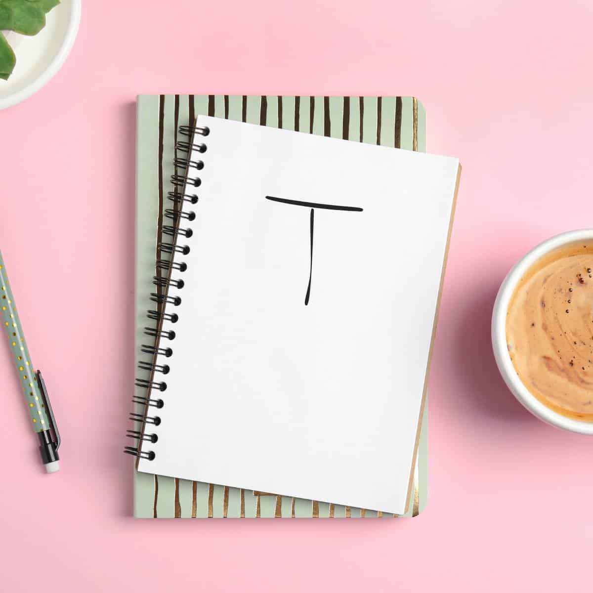 Flat lay photo of a handwritten half double crochet chart symbol on a notebook, accompanied by a mechanical pencil, and a cup containing a latte.