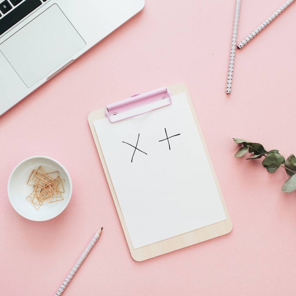 Flat lay photo of a handwritten single crochet chart symbol on paper clipped to a clipboard, with pencils, paperclips and a laptop nearby.