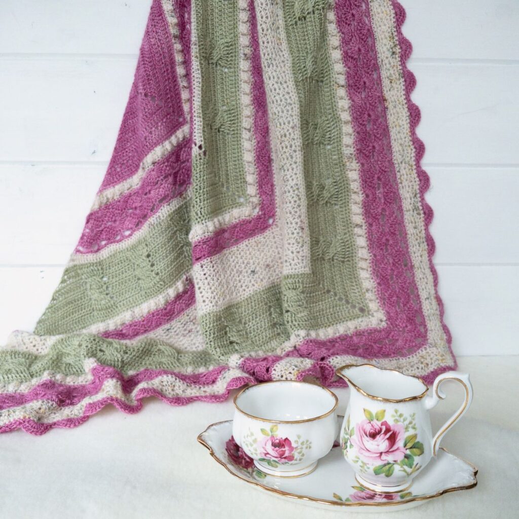 Triangular crochet shawl in soft pink, sage green, and cream stripes with textured stitch patterns and scalloped edging, draped behind a vintage floral teacup and creamer set.
