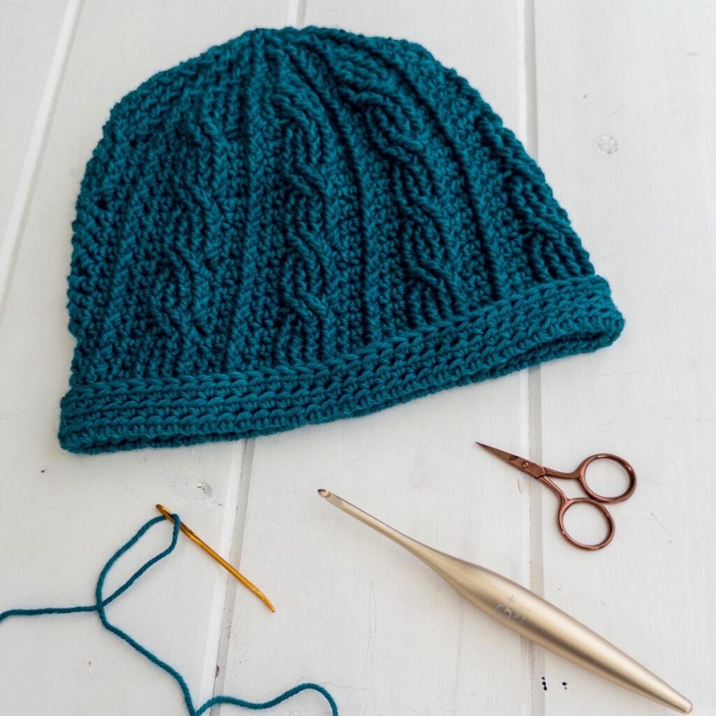 Teal knitted beanie hat with knitting needles and scissors on white background.