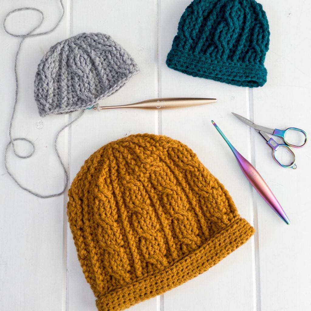 Layflat of three colors of The Sintra Cabled Crochet Hat showing textured cable stitches and double brim construction alongside crochet tools.