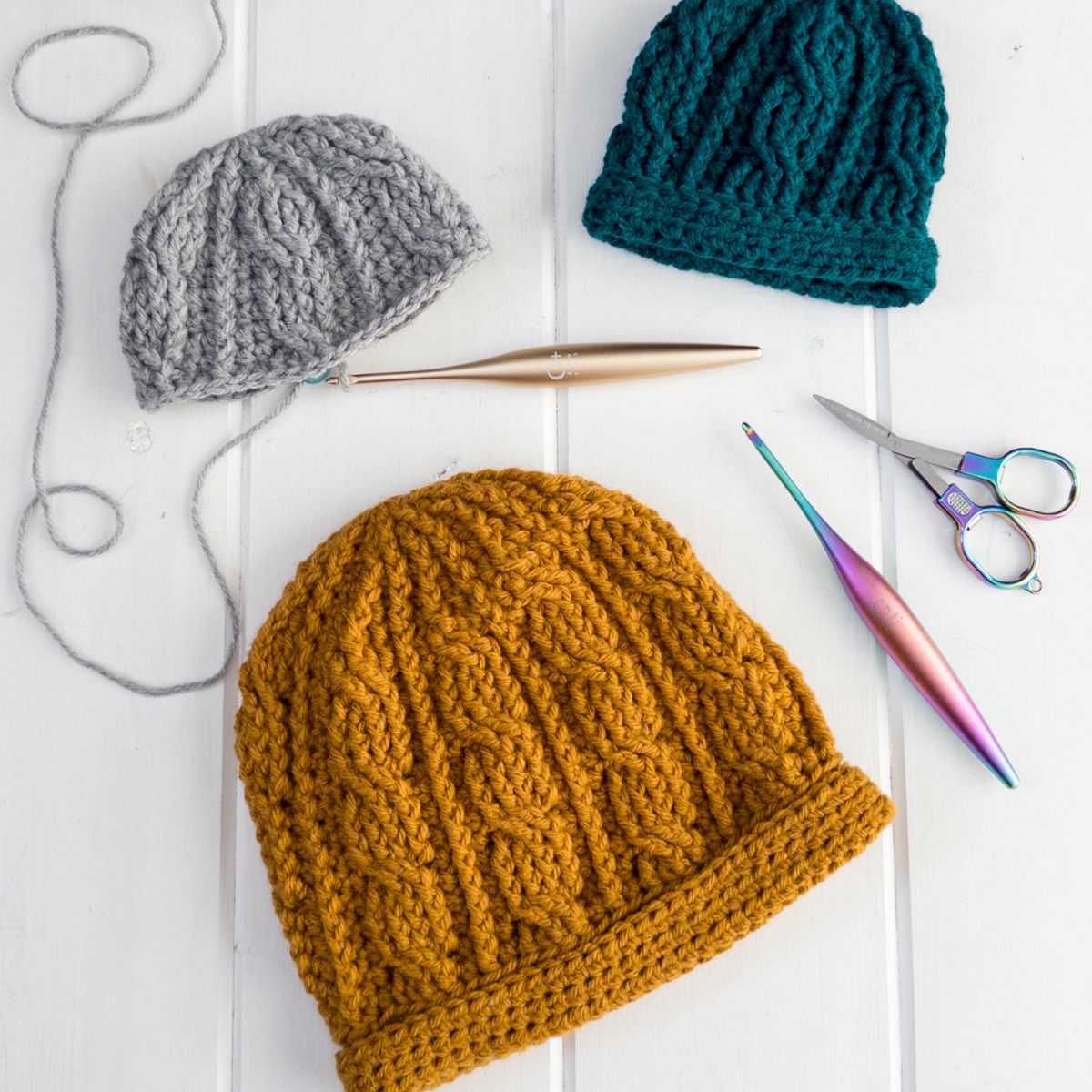 Layflat of three colors of The Sintra Cabled Crochet Hat showing textured cable stitches and double brim construction alongside crochet tools.